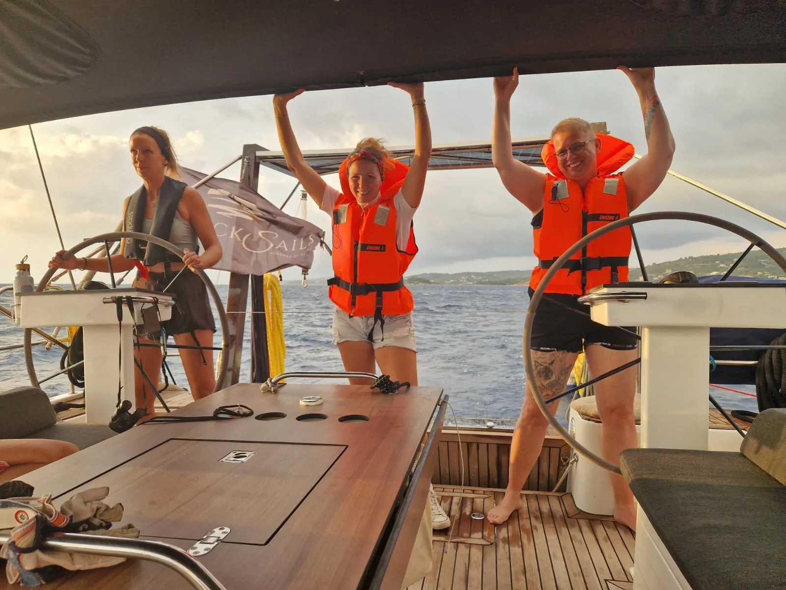 Drei Frauen im Cockpit unter Segeln: eine steuert, zwei halten sich in Rettungswesten lachend am Bimini fest